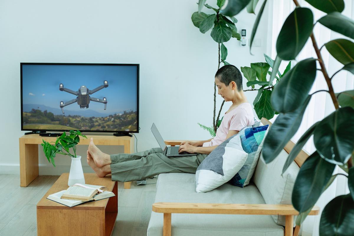 Man sitting on a couch with MacBook on his lap. TV displaying image of drone