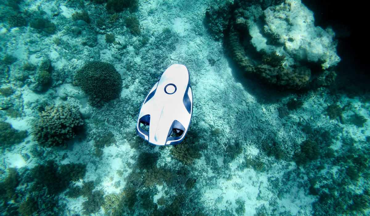 An overhead shot of a white underwater drone on water surface, among rocks and clear water