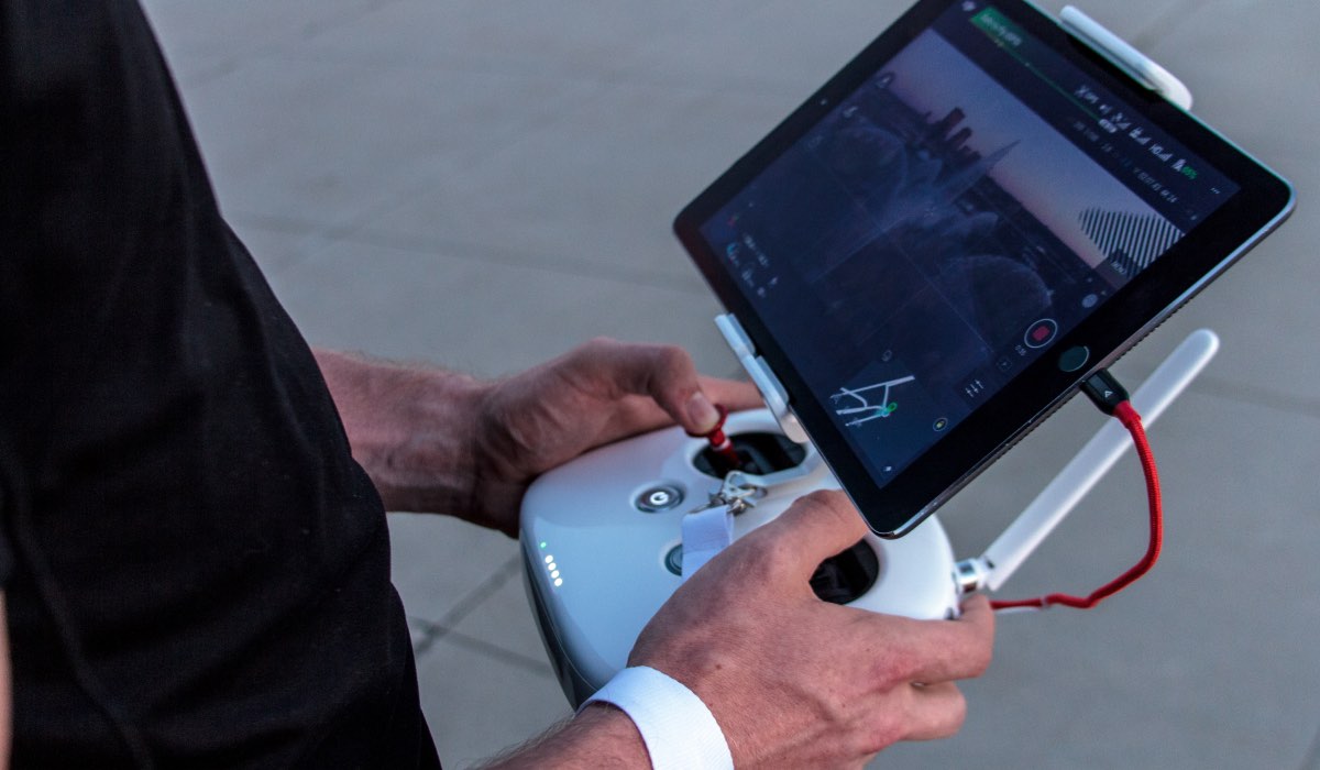 A person in a black t-shirt is holding a drone control console with a tablet showing drone cam feed
