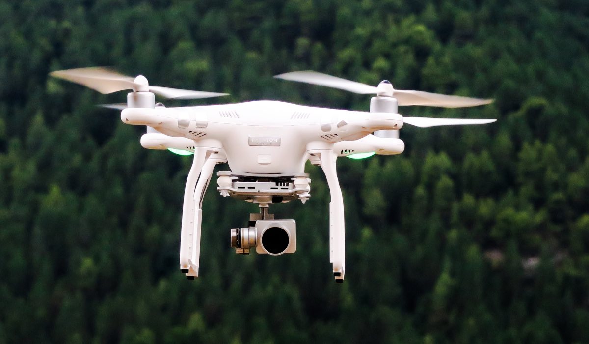 A white drone with a camera attached, flying above a forest